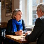 Deux professionnels européens d'une cinquantaine d'années en conversation intense dans un café, écharpe bleue vibrante sur la femme