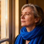 Femme d'âge mûr avec une écharpe bleue regardant pensivement par la fenêtre, évoquant une réflexion sur l'autisme adulte