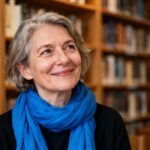 Femme d'âge mûr souriante avec un foulard bleu vif, assise dans une bibliothèque avec des étagères de livres en arrière-plan